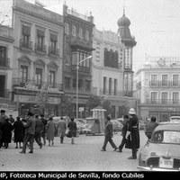 Aspecto de la Campana a principios de los años 60 del siglo XX. Instalado ya el Banco Hispano Americano en la esquina con O´Donnell, se conserva aunque remozado el edificio original de Aníbal González. Al fondo la Farmacia Central del doctor J.G. Espinar. Los viandantes y el escaso tráfico rodado se ven regidos por el guardia municipal. En el centro un motocarro con la cartelera de los cines del centro. ©ICAS-SAHP, Fototeca Municipal de Sevilla, fondo Cubiles